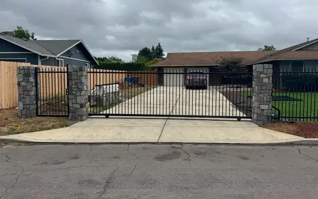 Black iron slide gate closed across concrete driveway between stone columns, ranch home — Interstate Gate Repair