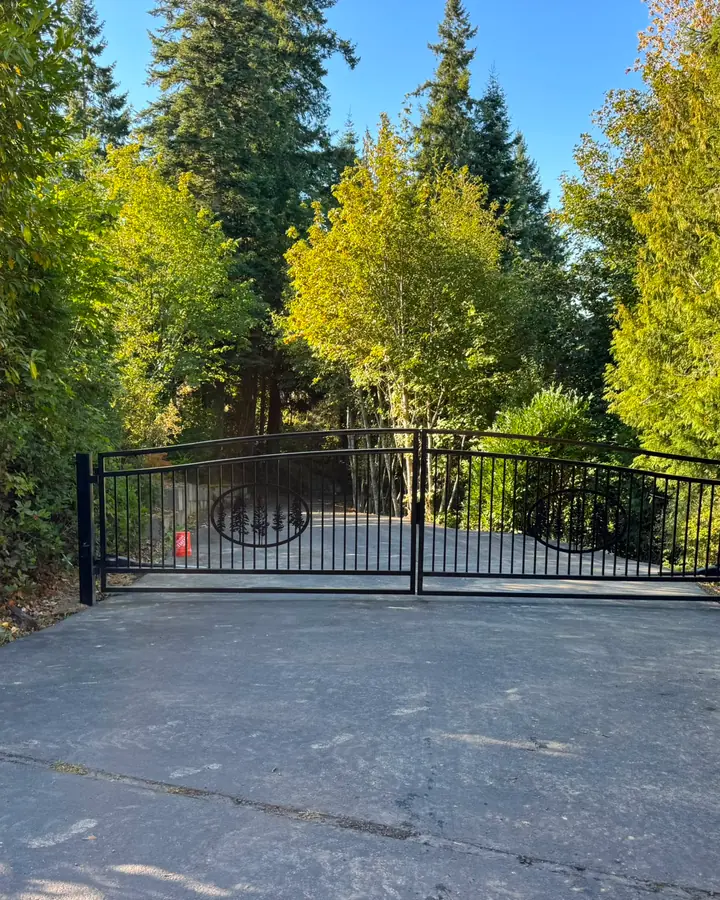 Black iron double swing driveway gate with tree motif and keypad, forested entrance — Interstate Gate Repair