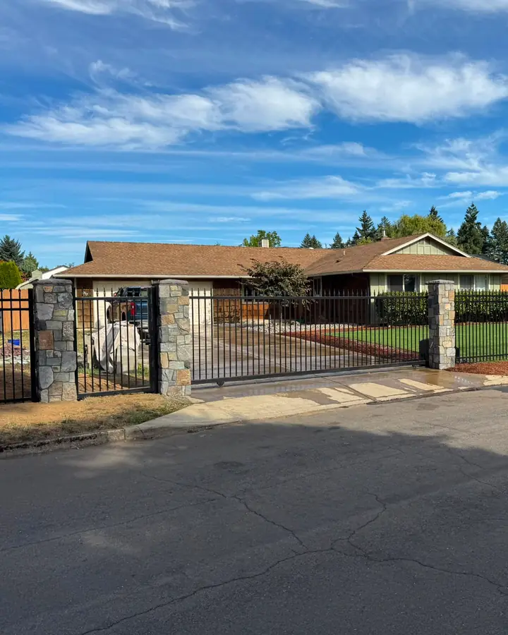 Wide street view of ranch home enclosed by black iron fence with stone columns — Interstate Gate Repair