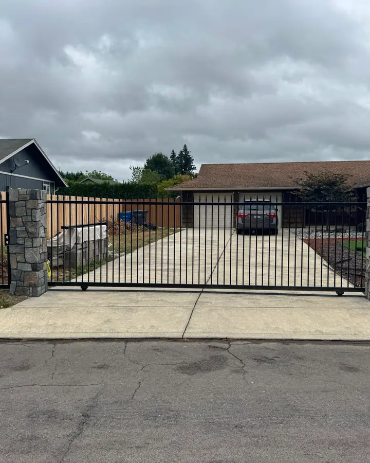 Black iron slide gate closed across concrete driveway between stone columns, ranch home — Interstate Gate Repair