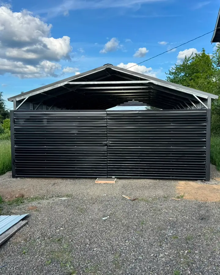 Black metal swing gate enclosing steel carport beside gray barn — Interstate Gate Repair