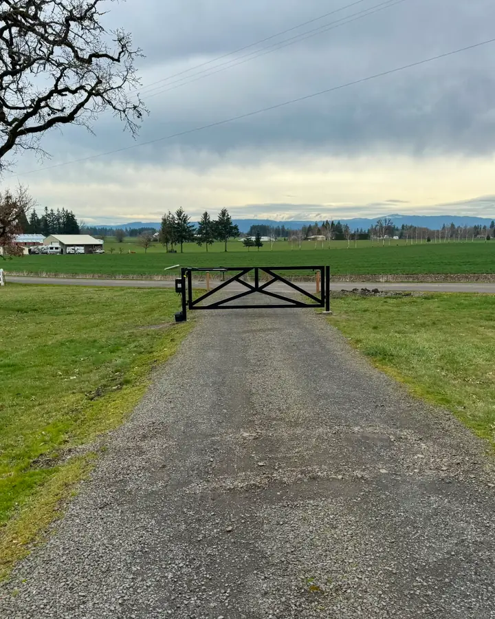 Wide view of closed black metal driveway gate across gravel drive with farm landscape — Interstate Gate Repair