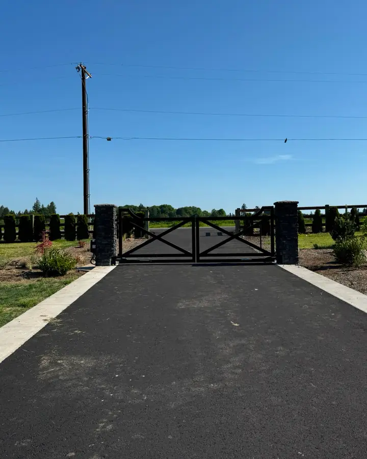 Black X-brace driveway gate with stone columns viewed across farm field — Interstate Gate Repair