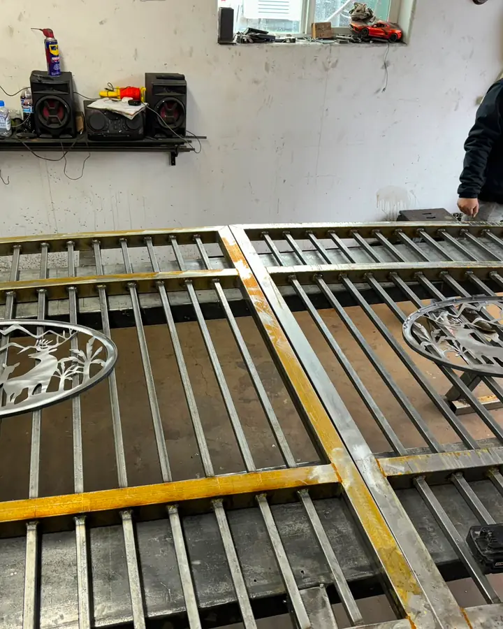 Two steel gate panels with deer scenic medallions on shop table, fabricator working — Interstate Gate Repair